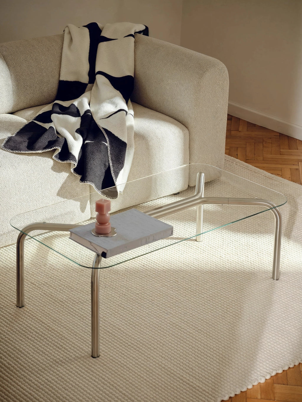 noo.ma Uku Coffee Table, a sleek rectangular glass table with silver metal legs, styled in a cozy living space. A cream textured sofa is draped with a black and white geometric patterned throw, and a pink candle sits on a book on the table.