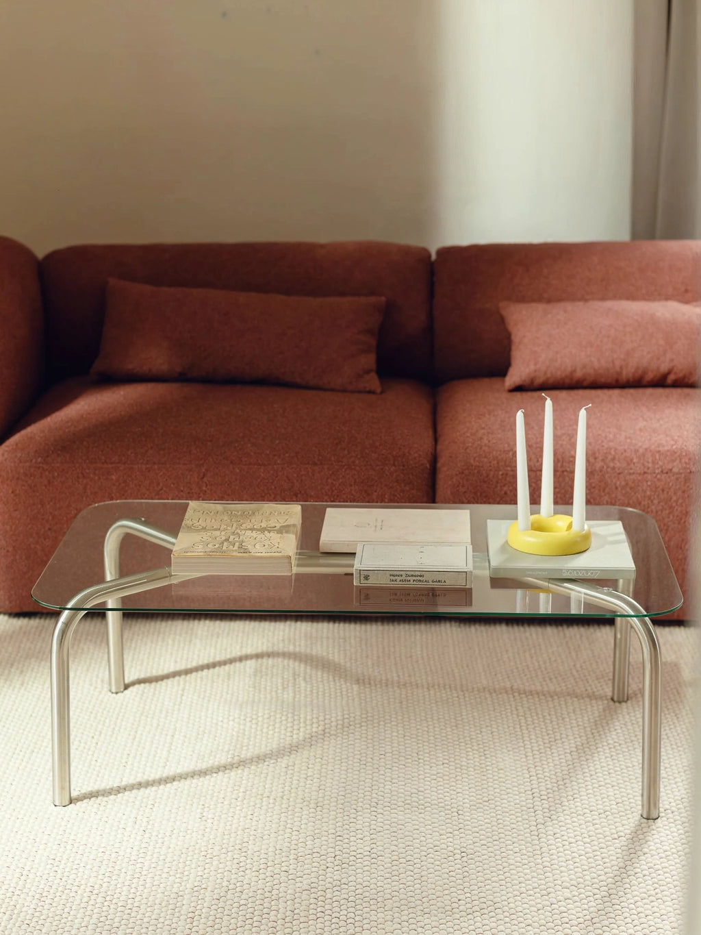 noo.ma Uku Coffee Table, a rectangular glass coffee table with elegant silver metal legs, placed in a stylish living room. A rich terracotta-colored sofa with matching pillows provides a warm backdrop, with books and a yellow candle holder on the table.