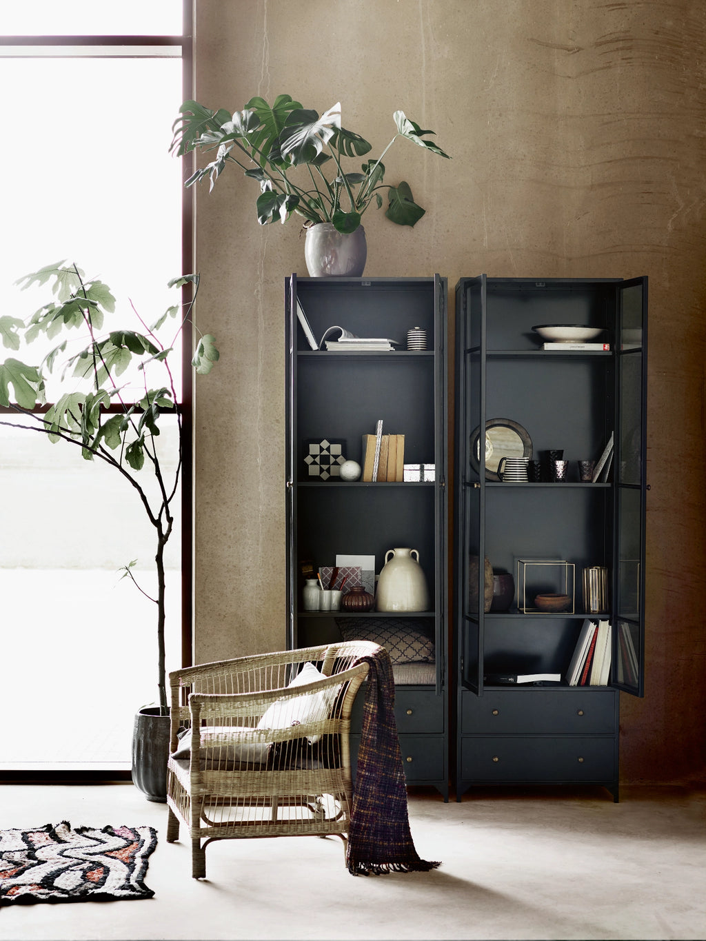 Tine K Metal High Cabinet, a sleek matte black industrial metal cabinet featuring two glass-paned doors with a grid design and two lower storage drawers with brass-toned pulls. This modern minimalist display cabinet offers versatile storage and a contemporary aesthetic for organized living. Perfect as a stylish bookcase for a living room, dining room, or industrial chic home office.