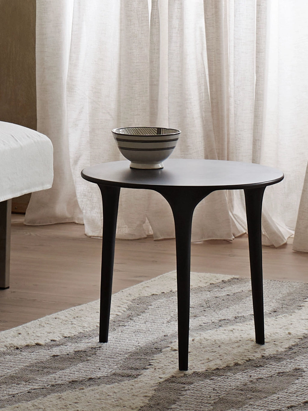 Tine K Feel Recycled Aluminium Table, a sleek round coffee table crafted from matte black recycled aluminium with a smooth finish and a minimalist aesthetic. It features a slender tabletop supported by three organically shaped, tapered legs that flare gracefully, embodying modern minimalist and Scandinavian design. This versatile black accent table is perfect for contemporary living rooms, cozy reading nooks, or as a chic bedside table in an organic modern bedroom.