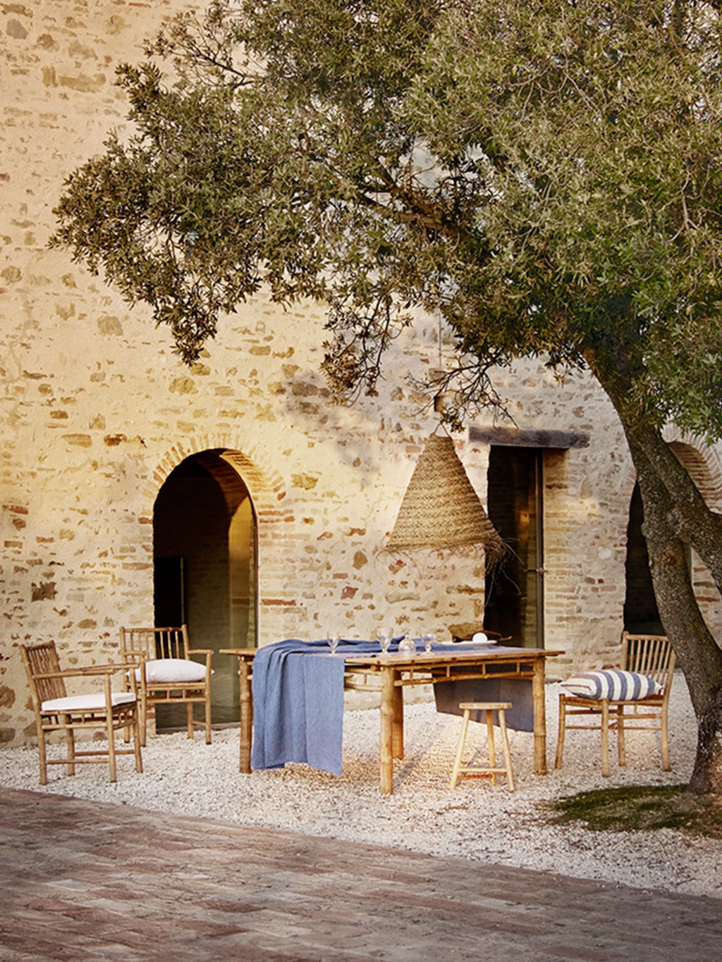 Tine K Outdoor Bamboo Dining Chair With Armrest - Set of 4, featuring a warm natural bamboo construction with a light brown finish, slatted backrest, and sturdy armrests, designed for comfortable al fresco dining. These rustic outdoor dining chairs include soft white linen cushions, perfect for creating an inviting Mediterranean or organic modern patio living space or garden setting.