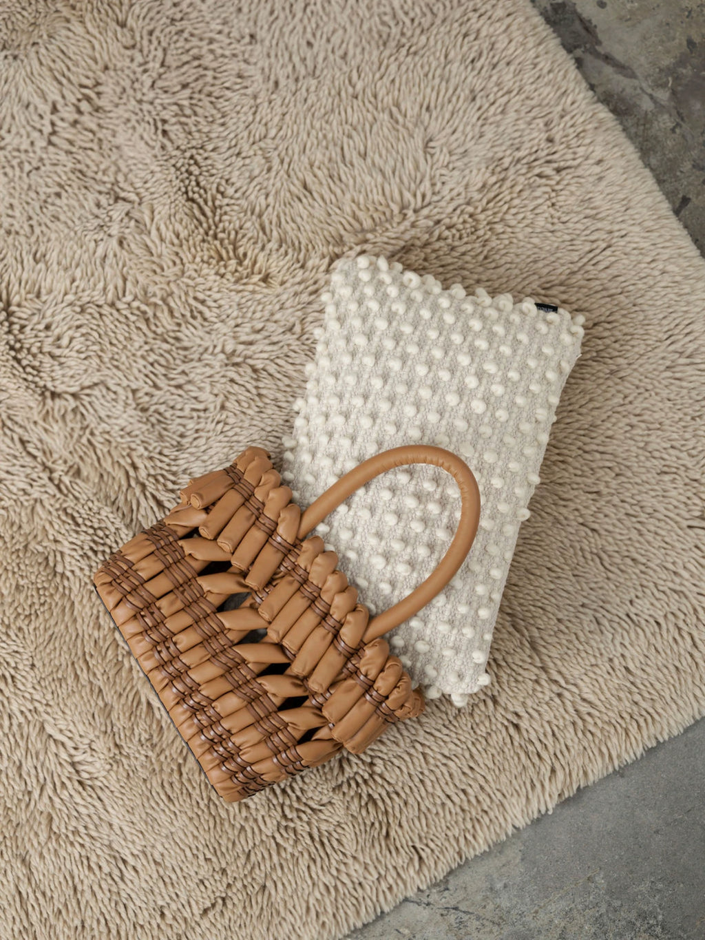 Tinted Objects Tuhlin Throw Pillow, a rectangular light beige cushion with a textured bobble knit pattern, resting on a plush, shaggy beige rug. This stylish decorative cushion adds cozy comfort to any modern living space.