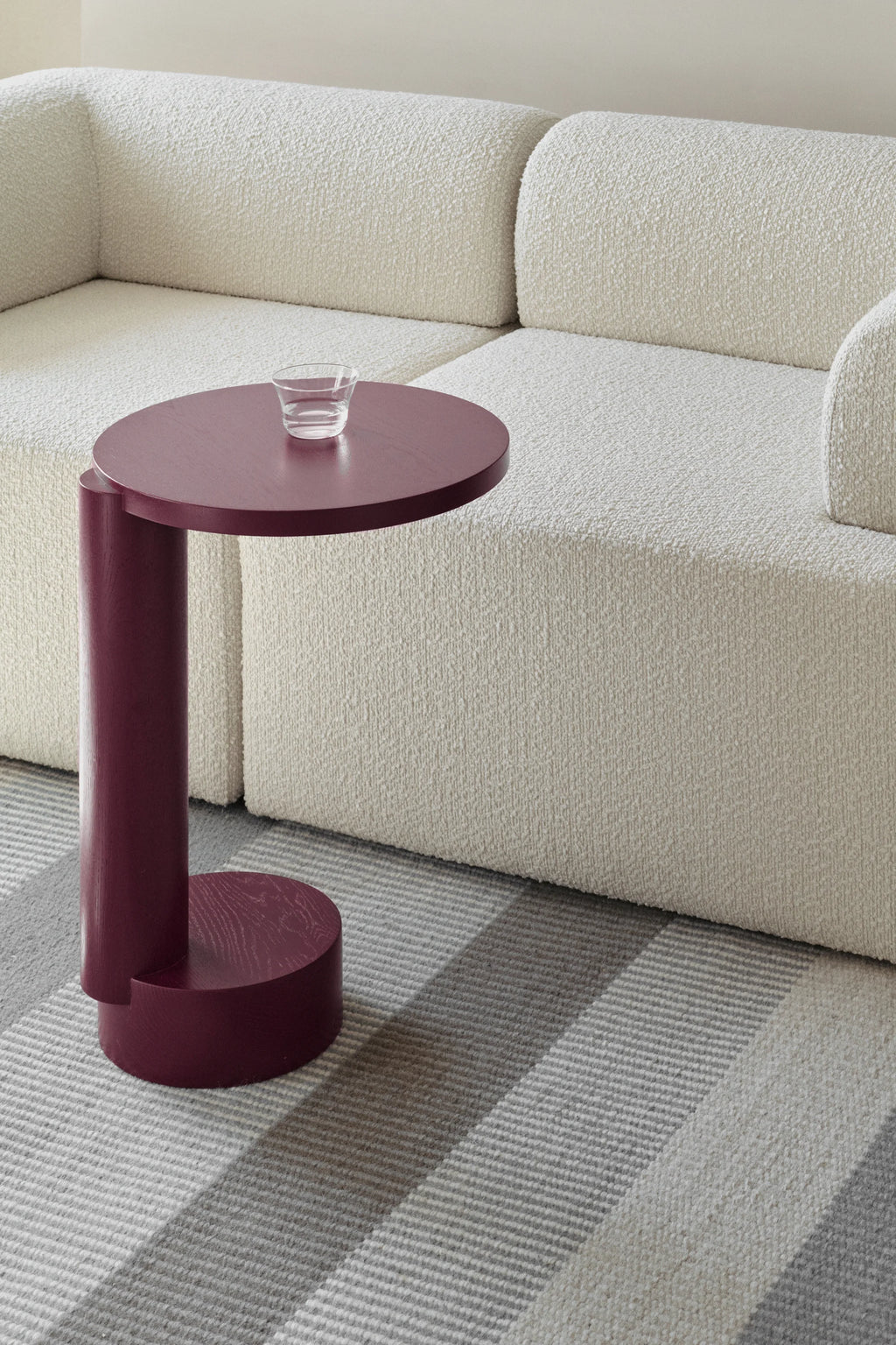 TAMO Bold Side Table, a premium matte merlot red wood C-shaped side table with wood grain visible, styled next to a textured cream sofa on a striped rug. Perfect for modern living spaces.