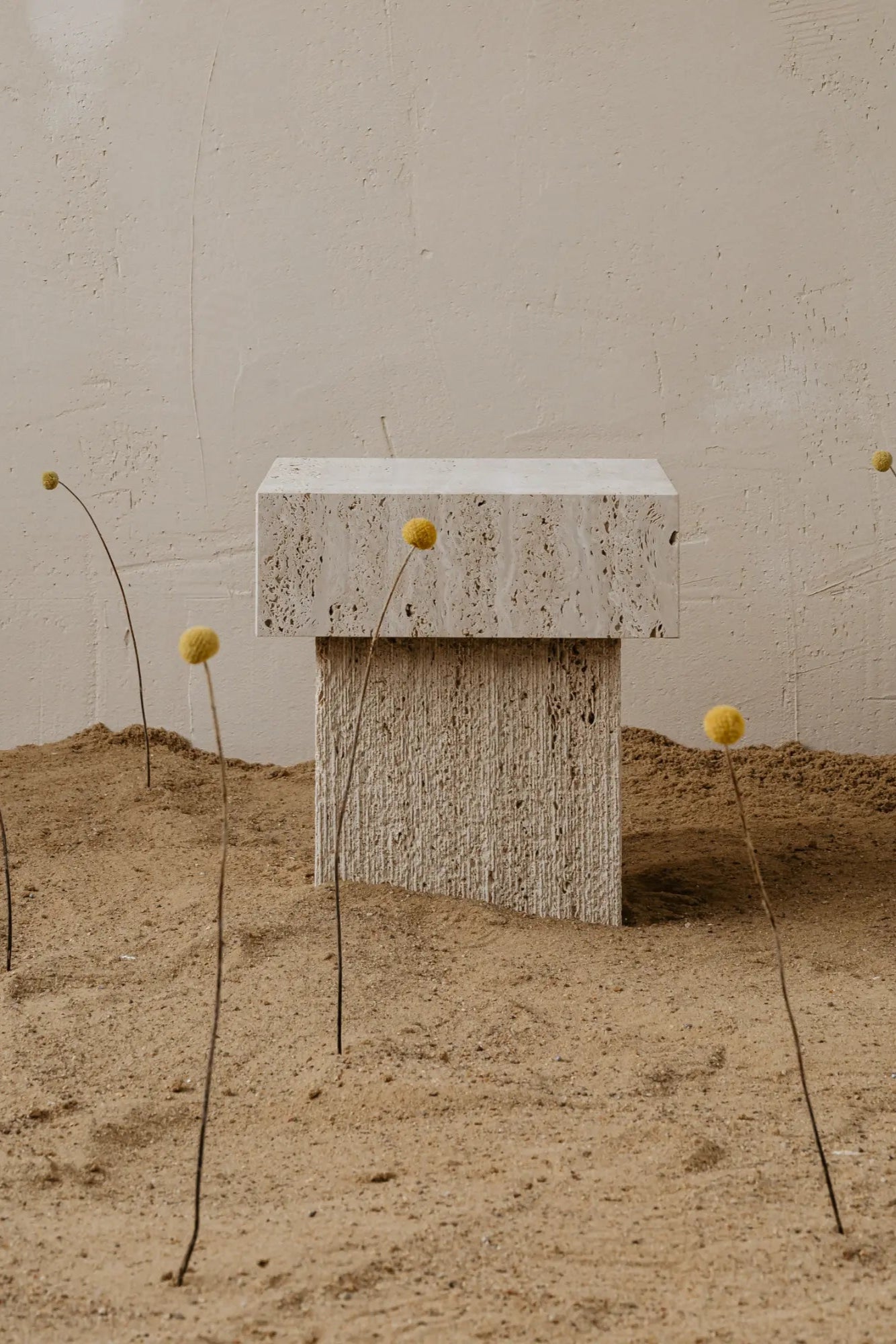 un'common Oro Side Table, a sculptural organic modern accent table crafted from natural beige travertine stone, featuring a porous rectangular top and a vertically ribbed, brutalist-inspired base. This minimalist travertine side table is perfect for a contemporary living room, bedroom, or as a striking display pedestal in Wabi-sabi or architectural interiors.