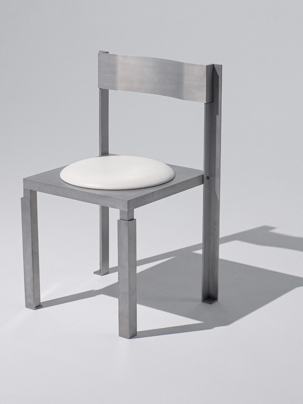 YET. Strut Chair, a striking industrial chic dining chair featuring a robust rectilinear frame of brushed silver-grey metal, likely stainless steel or aluminum, with distinctive strut-like legs and back support. It boasts a contrasting minimalist white upholstered circular seat pad, embodying a brutalist-inspired architectural design perfect for contemporary dining rooms or modern kitchen settings.