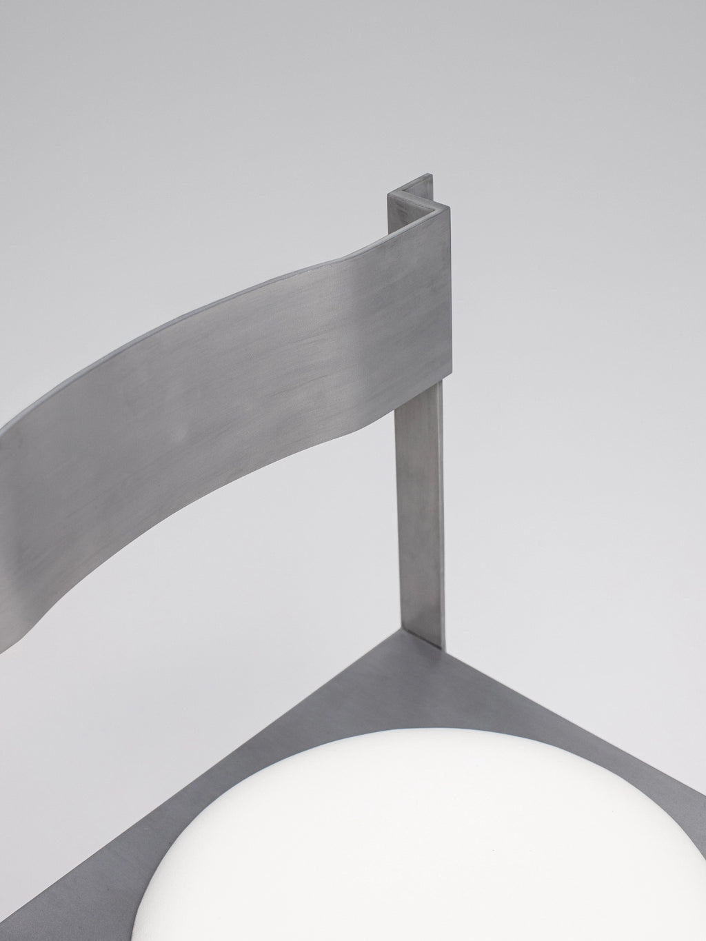 YET. Strut Chair, a modern minimalist dining chair crafted from brushed grey metal with a distinctive architectural, geometric frame and a subtly curved backrest. This contemporary steel chair features a contrasting round white seat, embodying an industrial chic design. Ideal for stylish dining rooms, modern kitchen furniture, or as an accent chair in minimalist interiors.