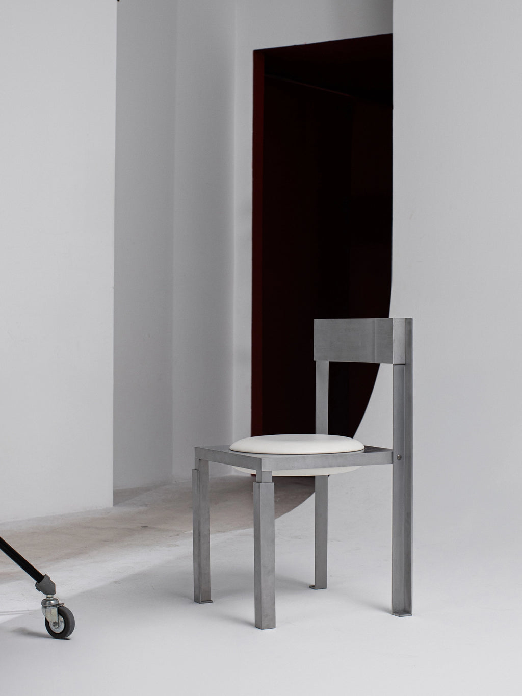 YET. Strut Chair, a brutalist-inspired dining chair featuring a robust matte grey metal frame with strong geometric lines and a minimalist aesthetic, complemented by a soft white upholstered circular seat cushion. Ideal as an industrial dining chair or contemporary accent piece for modern, architectural interiors.