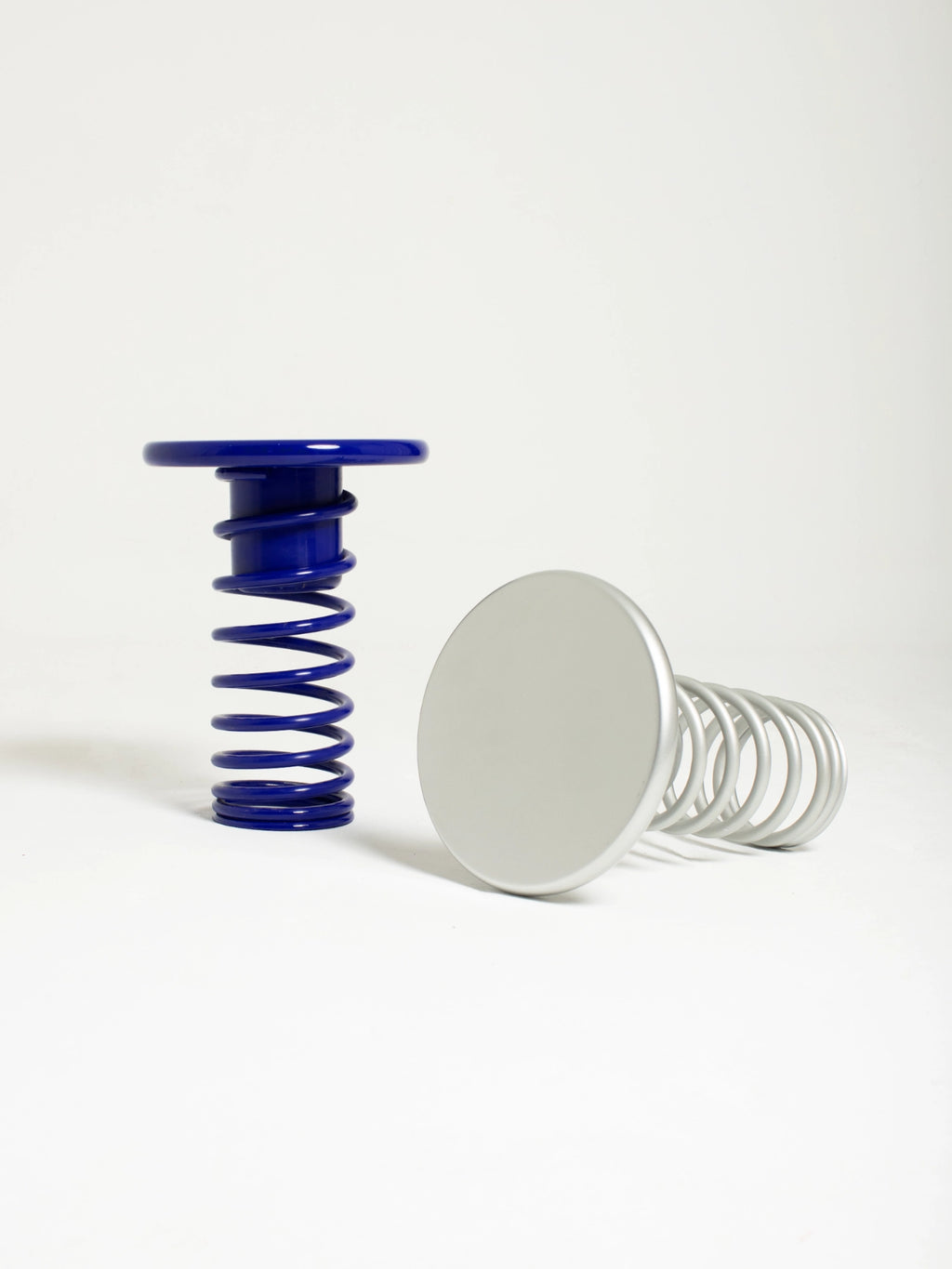 YET. Twist Stool, a playful postmodern accent piece featuring a bold royal blue lacquered metal spring base with a matching round top, alongside a matte silver version lying on its side, showcasing a unique sculptural design. This distinctive metal spring stool adds a vibrant, industrial-chic touch, perfect for a contemporary living room, creative studio, or playful bedroom accent.