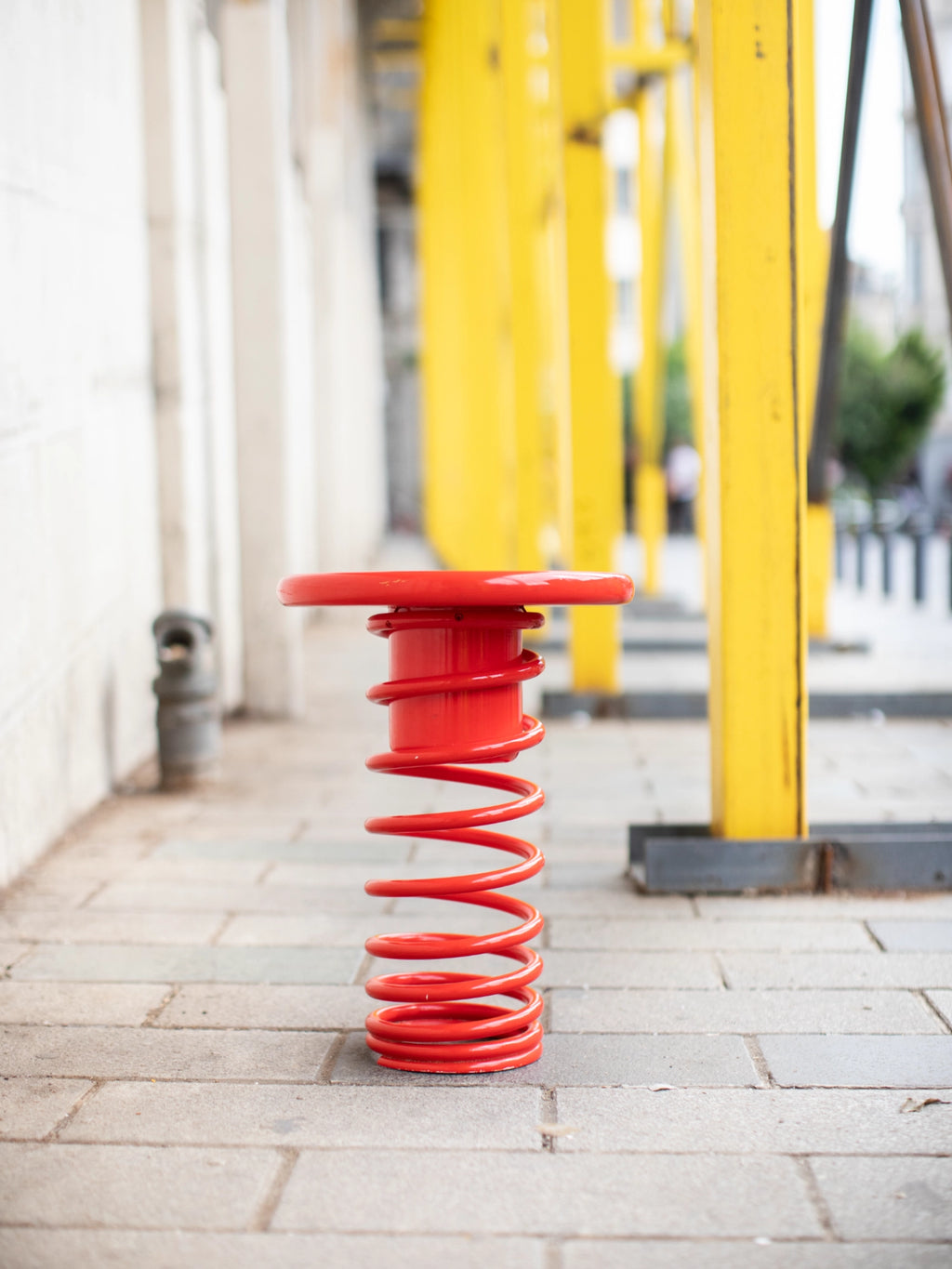 YET. Twist Stool, a vibrant red metal outdoor stool with a distinctive spiral spring base and a flat, round top, featuring a smooth, glossy finish. This modern industrial design stool offers playful yet durable public seating for urban landscapes and contemporary outdoor installations.