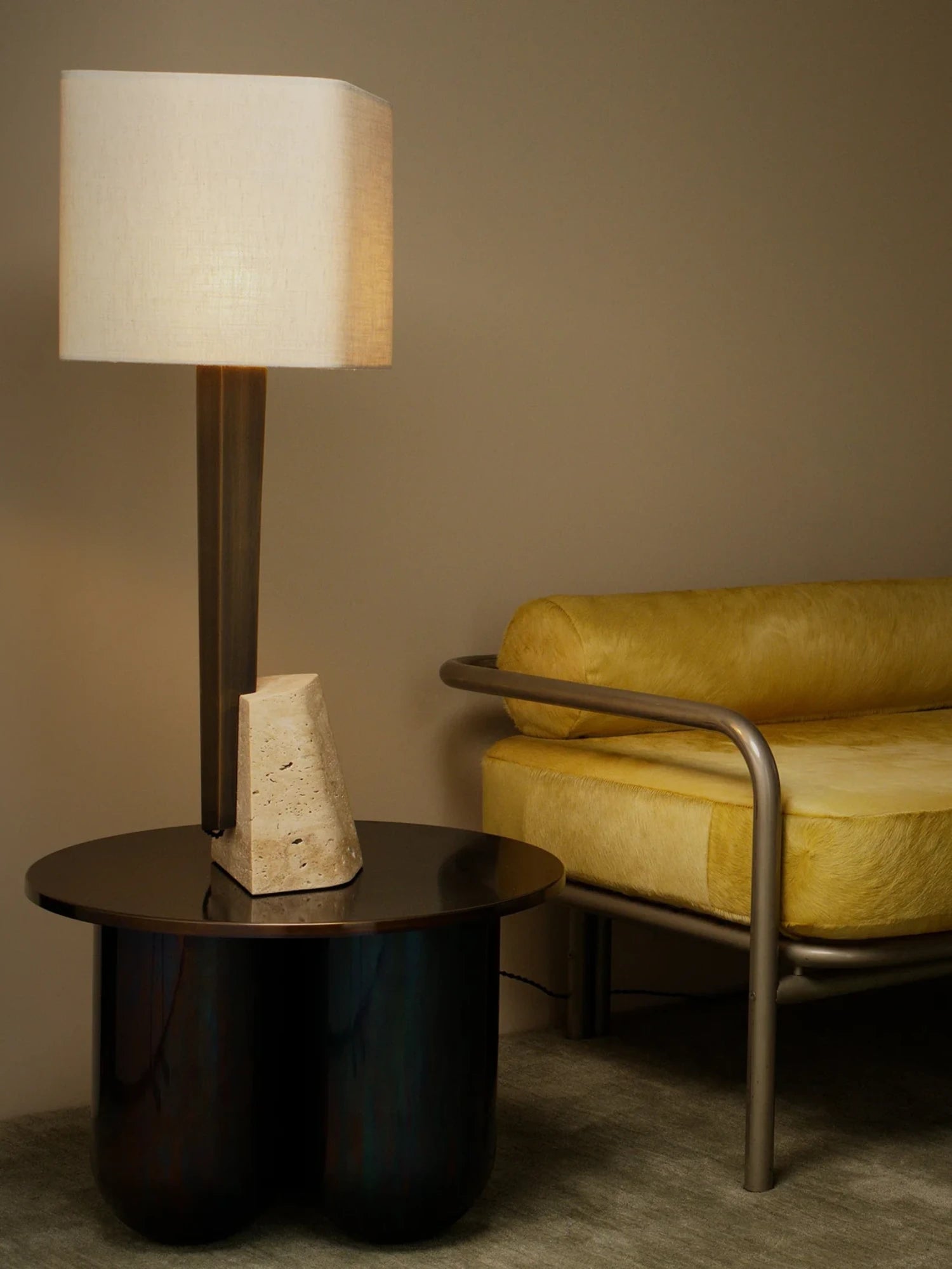 A large, modern Roc Table Lamp with a square, off-white lampshade is on a round, dark side table with three thick legs. A blocky, textured concrete or stone object is next to the lamp's base. To the right, a long, low daybed with a metal frame and yellow, fuzzy cushions is positioned against a gray wall. A small black pillow is on the daybed.